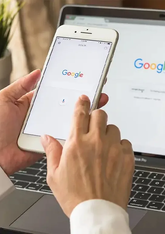 Personne utilisant la recherche vocale Google sur un smartphone, avec la même page affichée sur un ordinateur portable en arrière-plan.